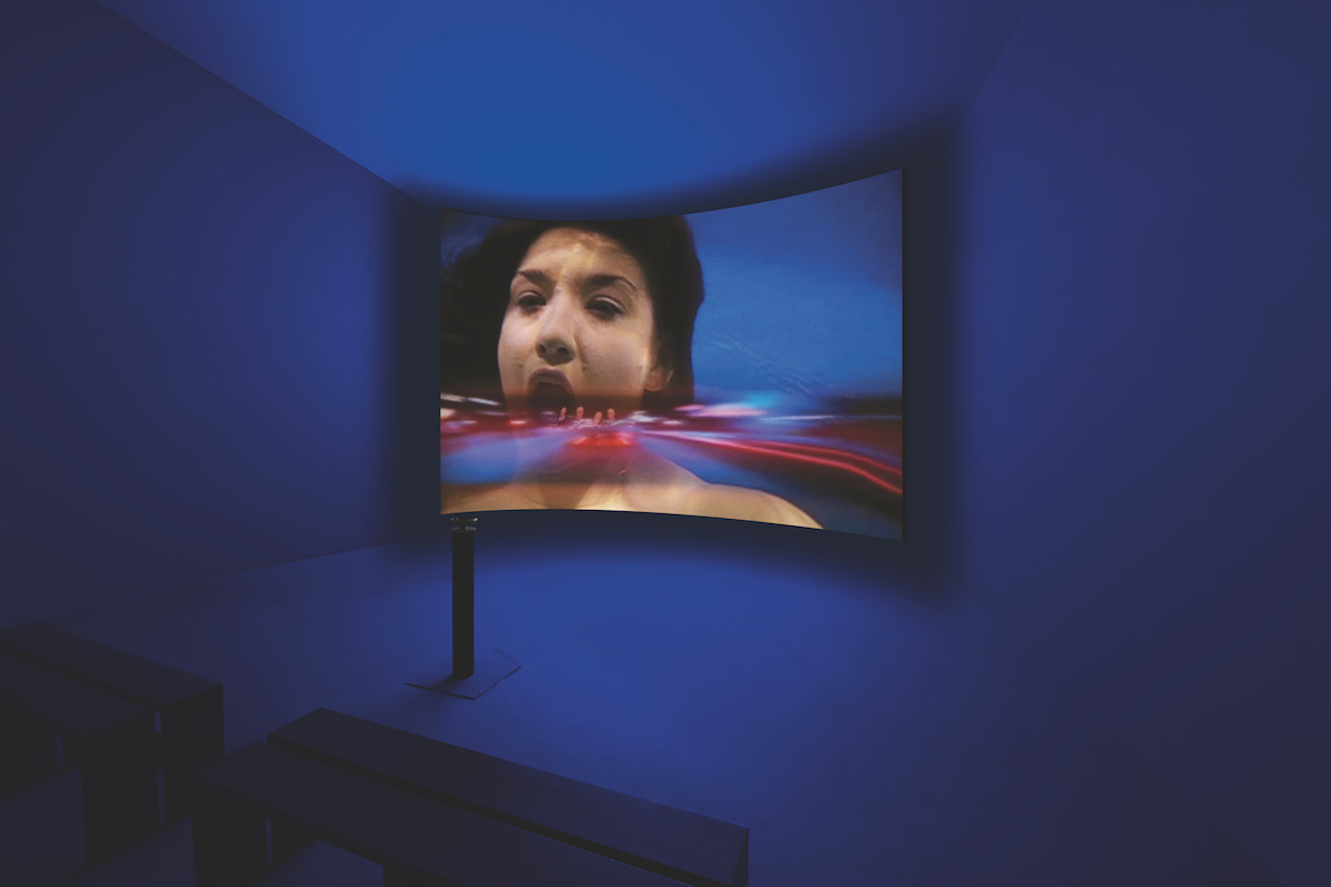 In a darkened room, a giant curved screen shows a white woman opening her mouth. A blur of red and blue appears to recede into it.