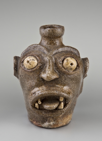 Ceramic vessel in the shape of a head with bulging eyes and a gaping mouth with four teeth and tongue visible.