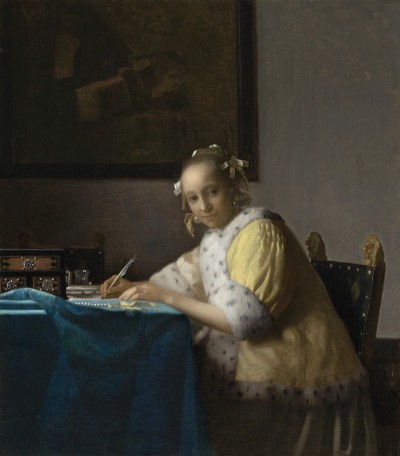 A white woman in a yellow jacket with a furred collar sits at a table writing a letter. The table is covered in a blue cloth. She looks out at the viewer. Behind her is a painting that cannot be made out.