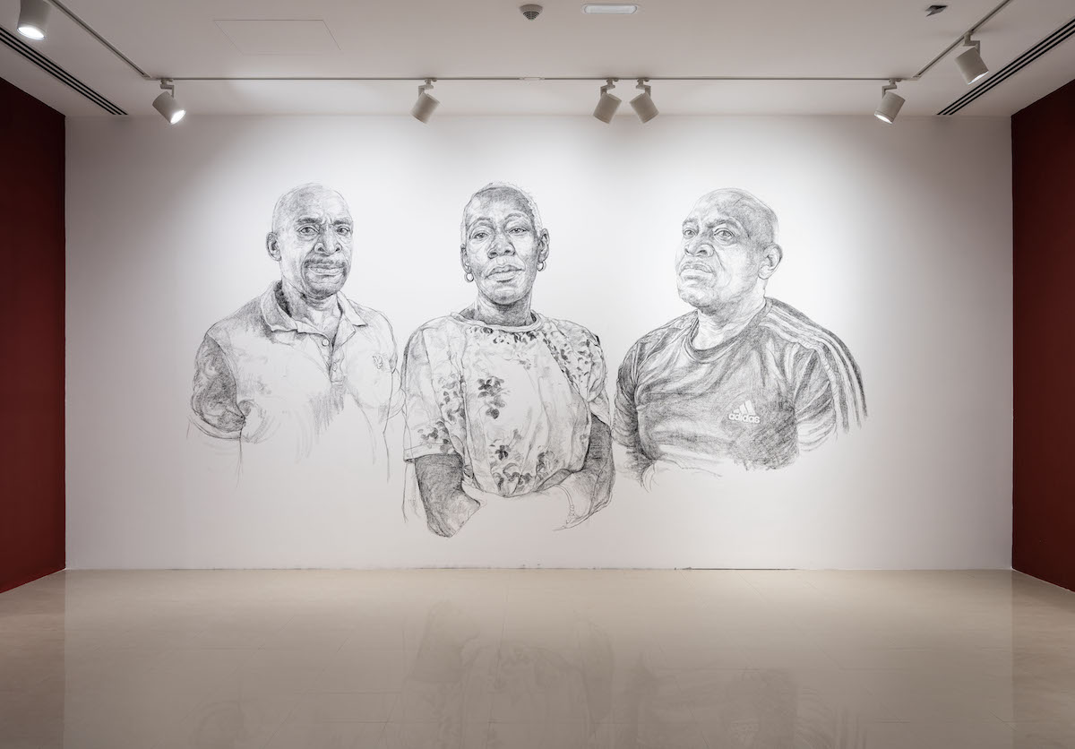 A white gallery wall with three portraits of Black people drawn on it. They resemble a man in a polo, a woman in a dress, and a man in a T-shirt.