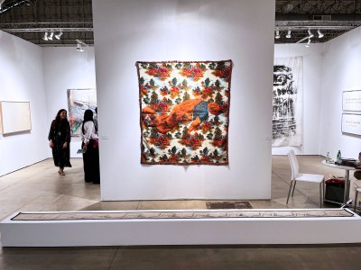 View of an art fair booth showing a bedspread painted with a man's body that is hung on a wall, with a long, scroll-like work on a short plinth in front.