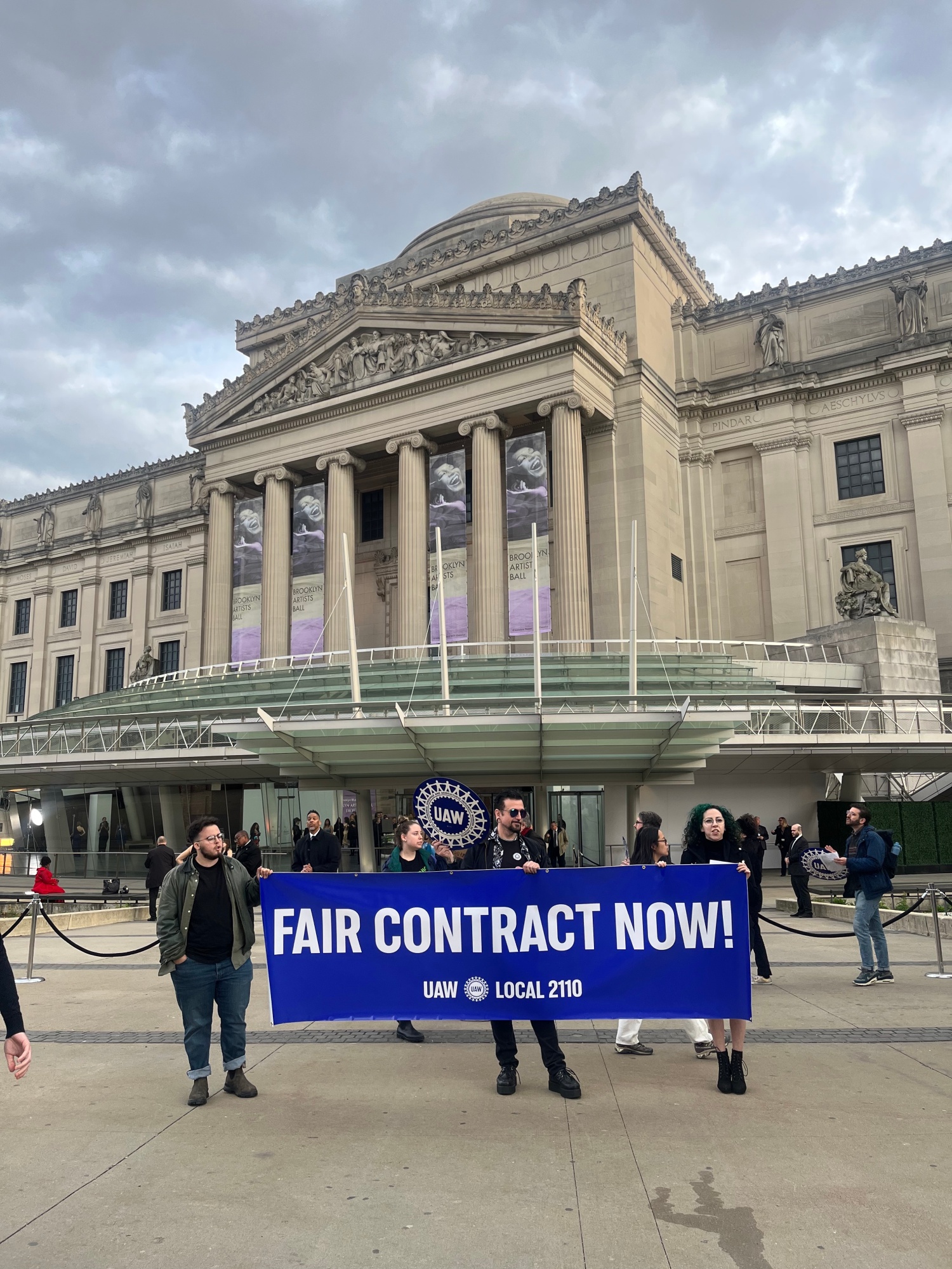 People hold a blue sign that reads 'FAIR CONTRACT NOW' in front of the Brooklyn Museum building.