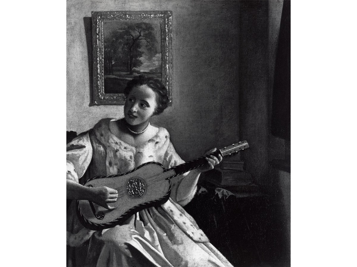 A black-and-white image of a white woman strumming a guitar beneath a painting of a tree.