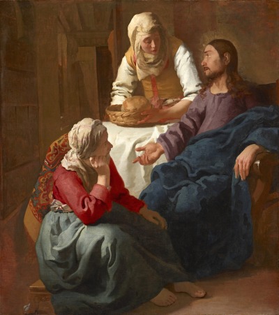 Two white women and a white Jesus Christ inside a nondescript room mainly lit in shits of brown. Jesus Christ, wearing a blue robe and a purple undershirt, points down to a seated woman, who wears a green dress and looks up at him admiringly. A standing woman behind him wearing brown places a basket with a loaf of bread in it on a table laid with a cloth.