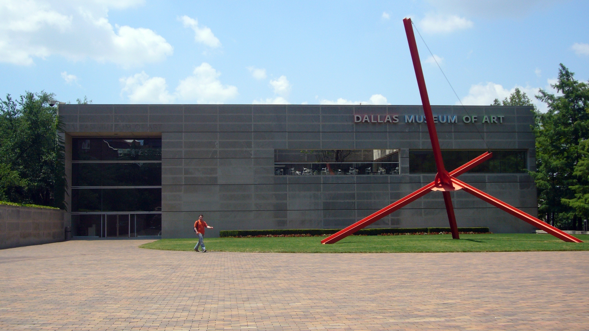 An angular red sculpture stands outside a rectangular building.