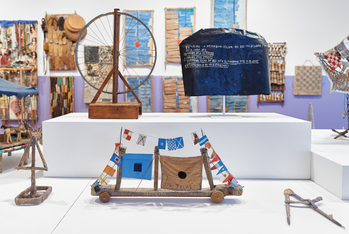 A wheel, a boat-like assemblage, and a piece of tarp with Portugese text are in the foreground. The wall in the background is brimming with out-of-focus-assemblages.