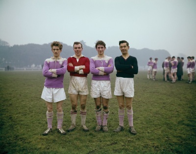 four male rugby player standing side by side in a grassy field, wearing white shorts, knee socks, and purple, red, or black tops