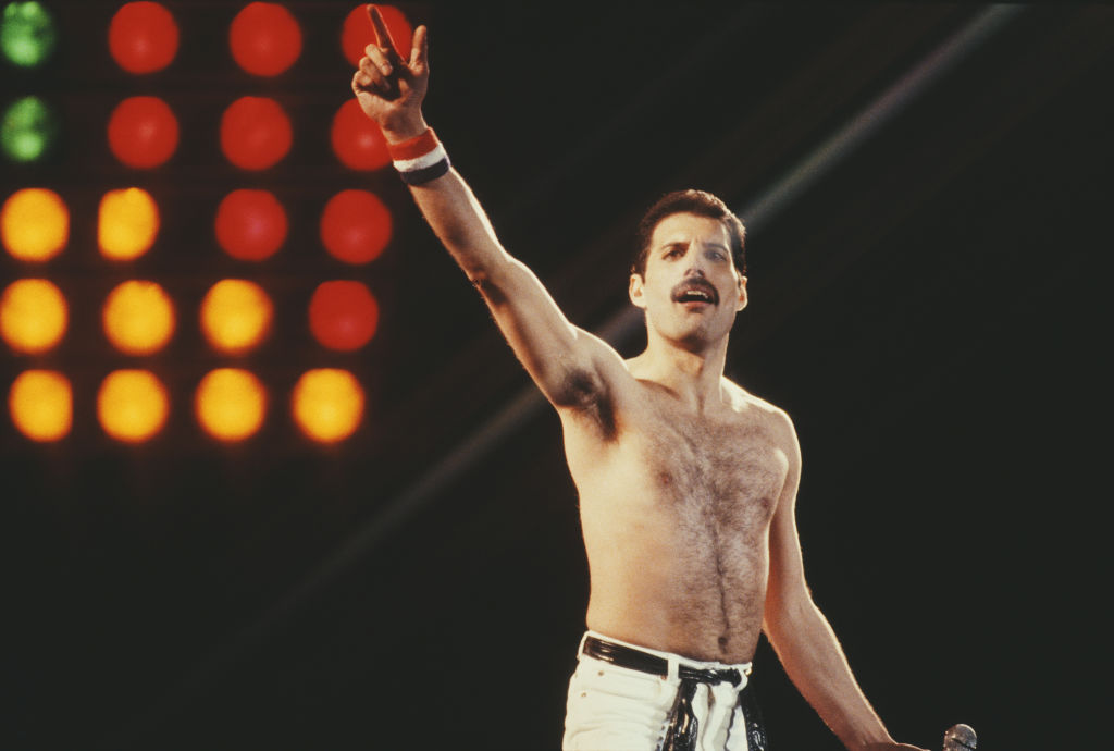 A shirtless man points a finger to the heavens. He holds a microphone in his other hand, aarkness and a few lights loom behind him.