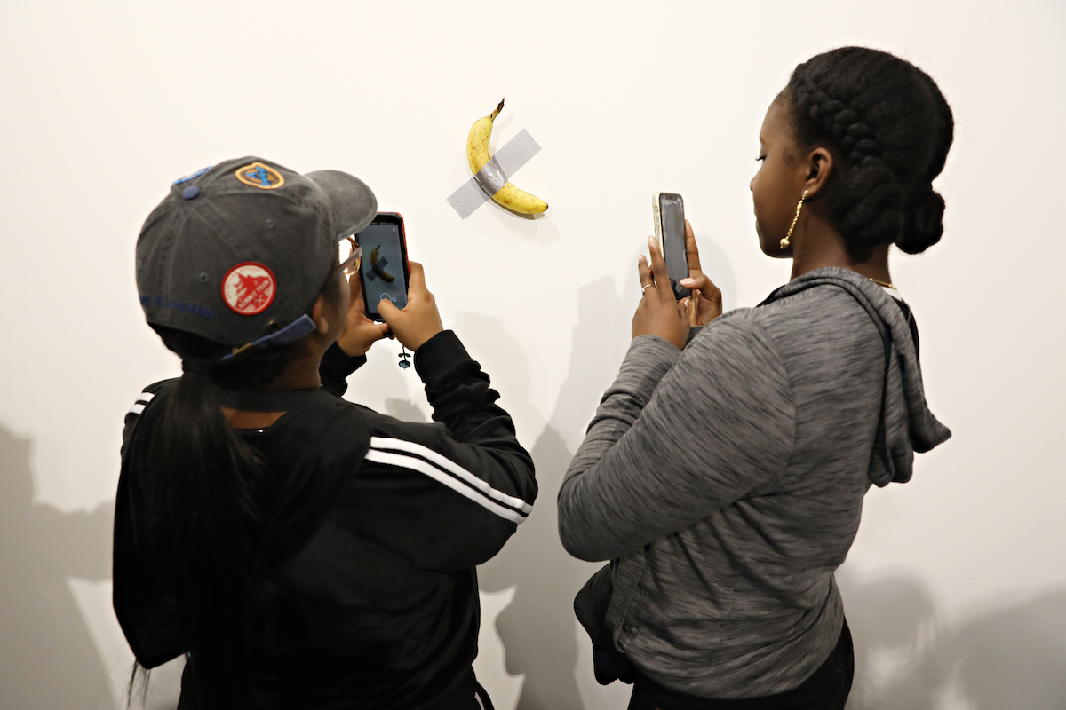 Two young Black women in baseball hats taking photos of a banana duct-taped to a wall.