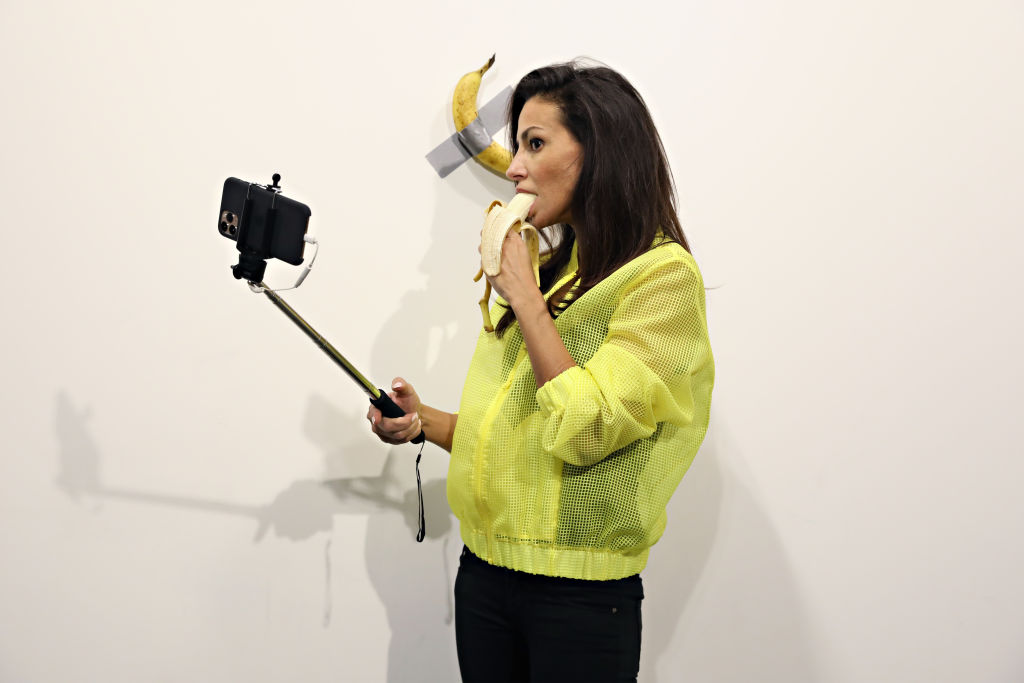 A woman eats a banana and poses for a selfie (with the aid of a selfie stick) in front of a banana that is taped to a white wall with gray tape.