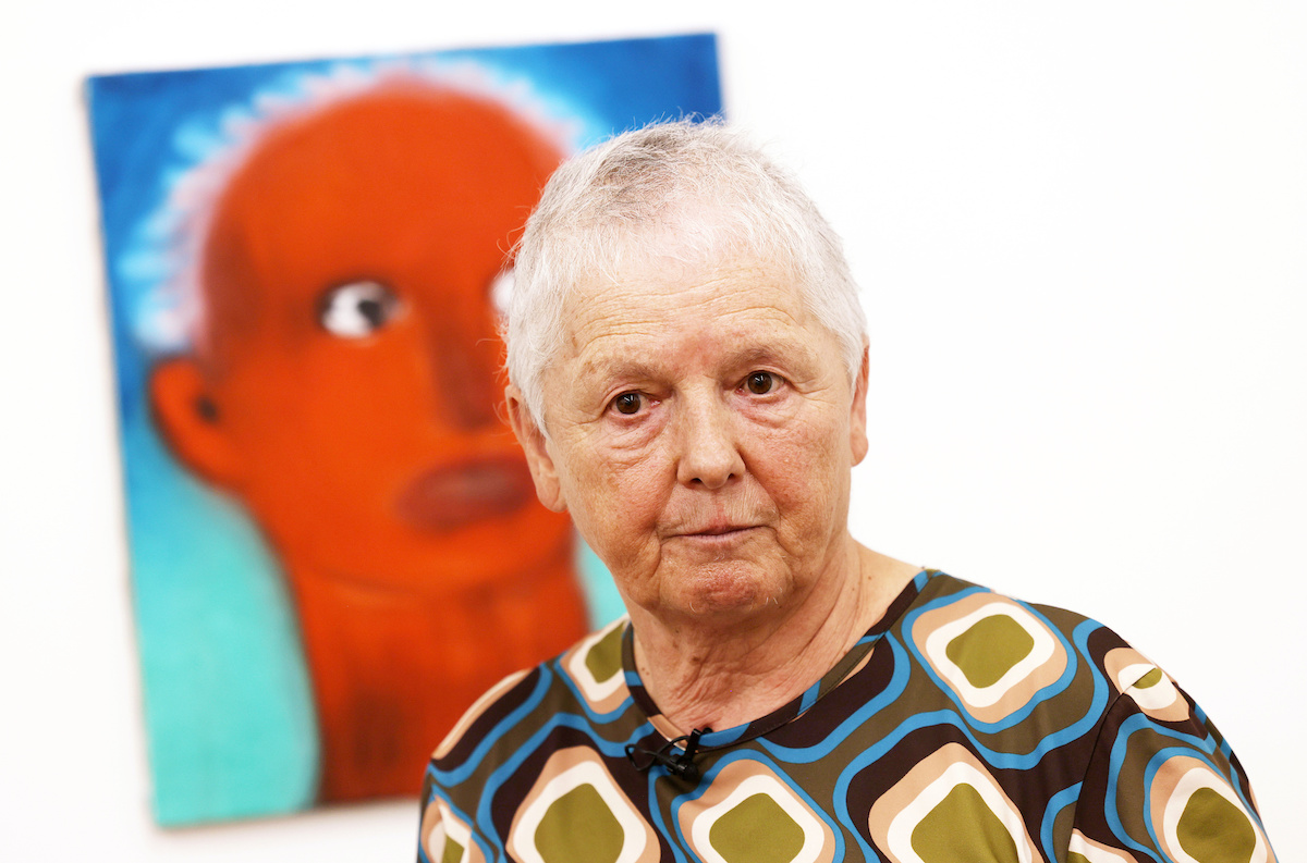 A stone-faced woman standing in front of a painting of a bright red face similar to her own.