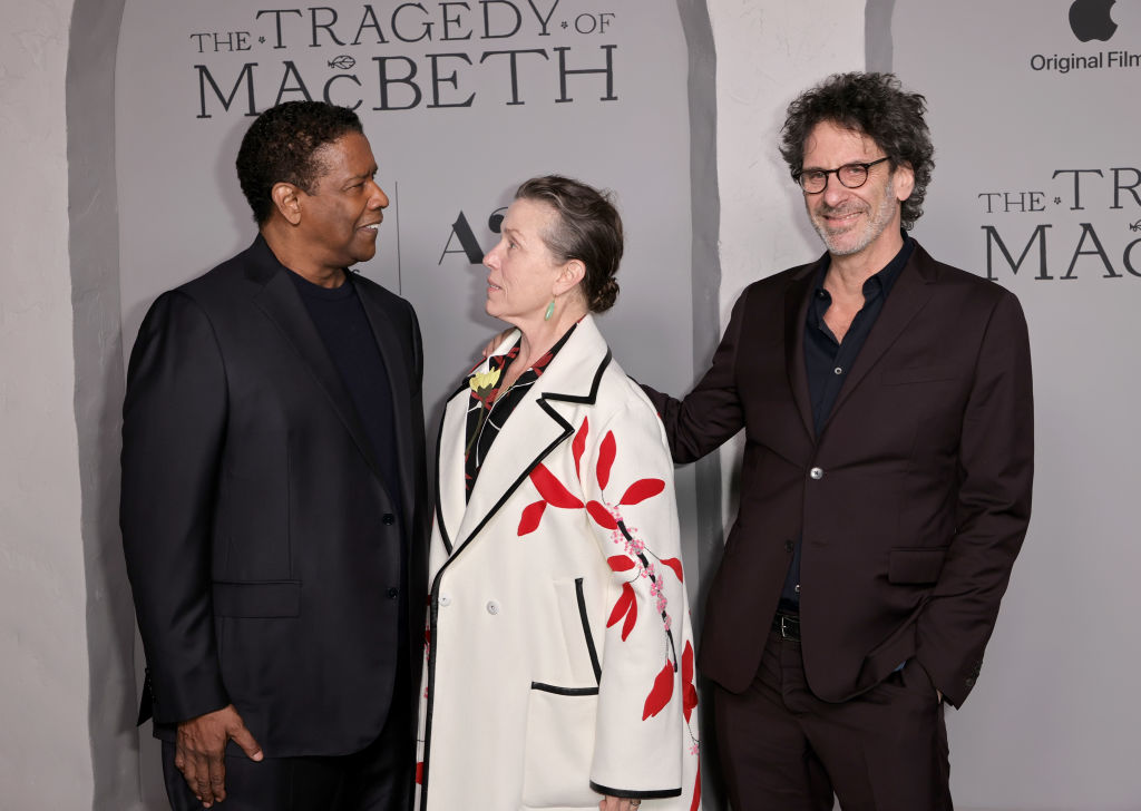 A man, a woman, and a man stand together in front of a sign that says "The Tragedy of Macbeth." The first two look at one another.
