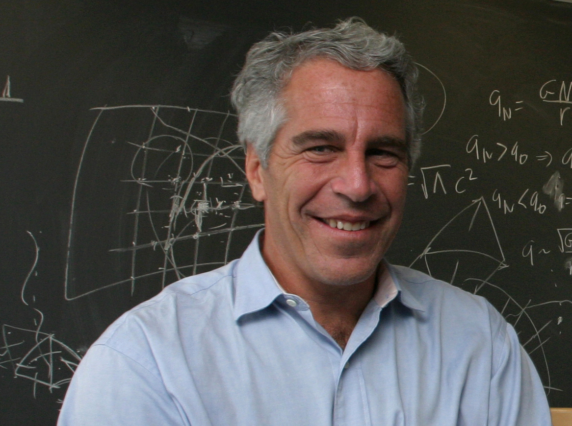 A white man with salt and pepper hair smiling in a blue shirt. He stands in front of a blackboard with mathematical equations scrawled on it.