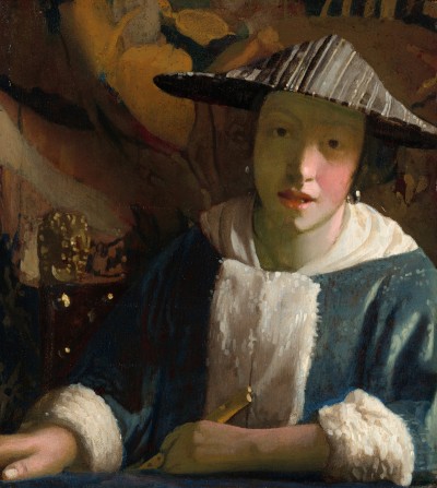 A white woman sitting at a table wearing a blue shirt adorned with white furs. She holds a flute in one hand that is leaned against the table. Behind her is a chair and a large painting.