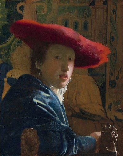 A young white woman seated in a chair pictured from her waist up. She has turned herself around to face the viewer, placing one arm the back of her chair. She wears a deep blue shirt that has white ruffles and a bright red hat. Behind her is a tapestry that depicts a building.