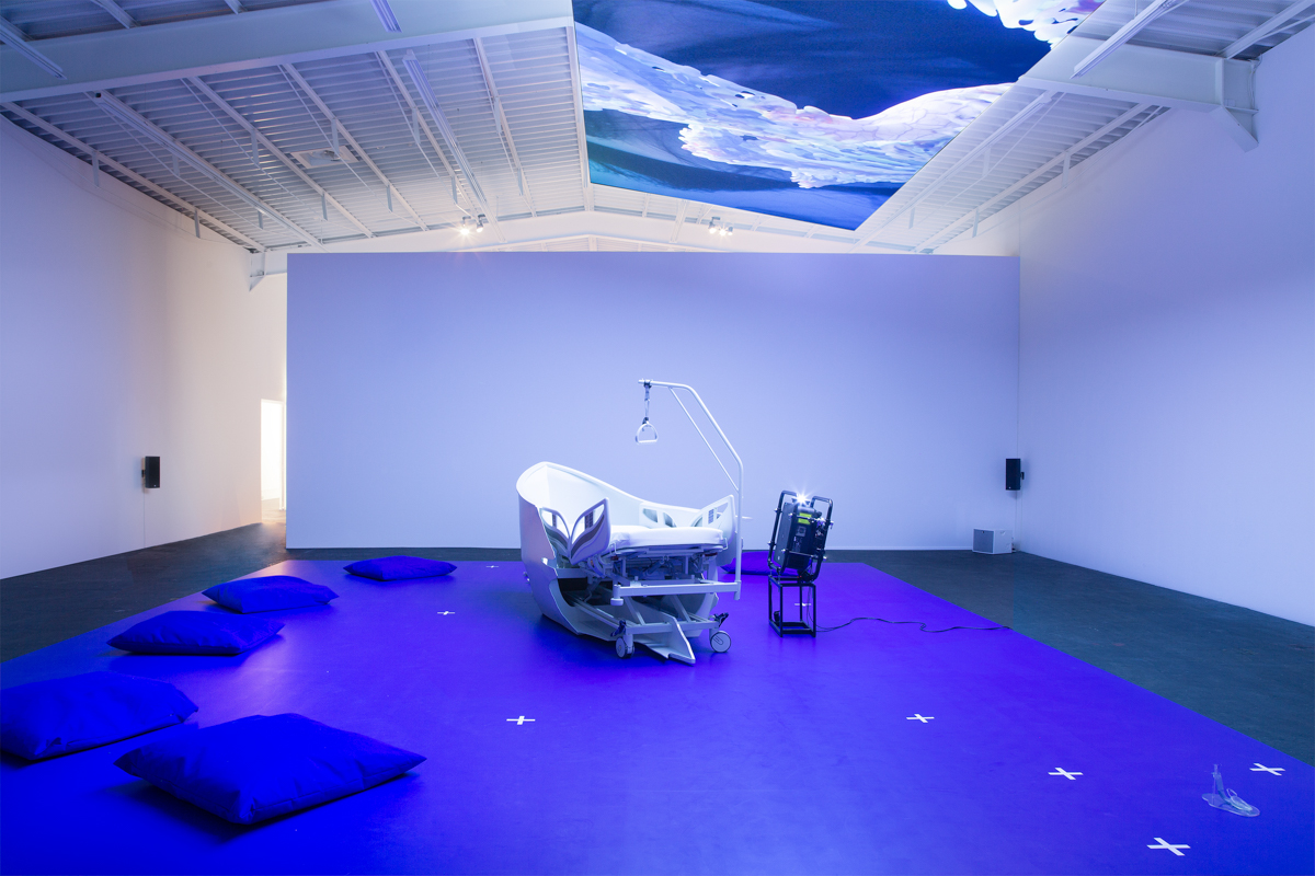 Blue cushions and a hospital-bed-cum-boat sit atop a big blue plinth in a gallery. There is a projection pointed toward the ceiling creating a blue image that is hard to make out.