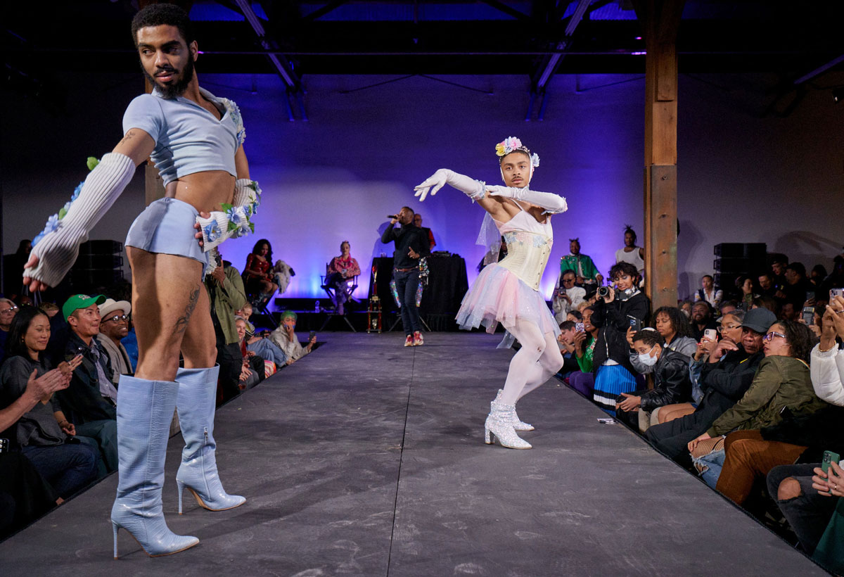 Two Black queer performers walk a runway as part of a ball.