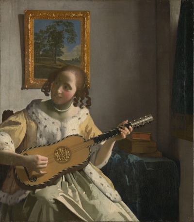 A smiling young white woman strumming a guitar. She wears a yellow dress with furs, and her hair is plaited with curls. Behind her is a partly visible table with a pile of books. A painting hanging on a wall above her head shows a quaint countryside scene dominated by a tall green tree.
