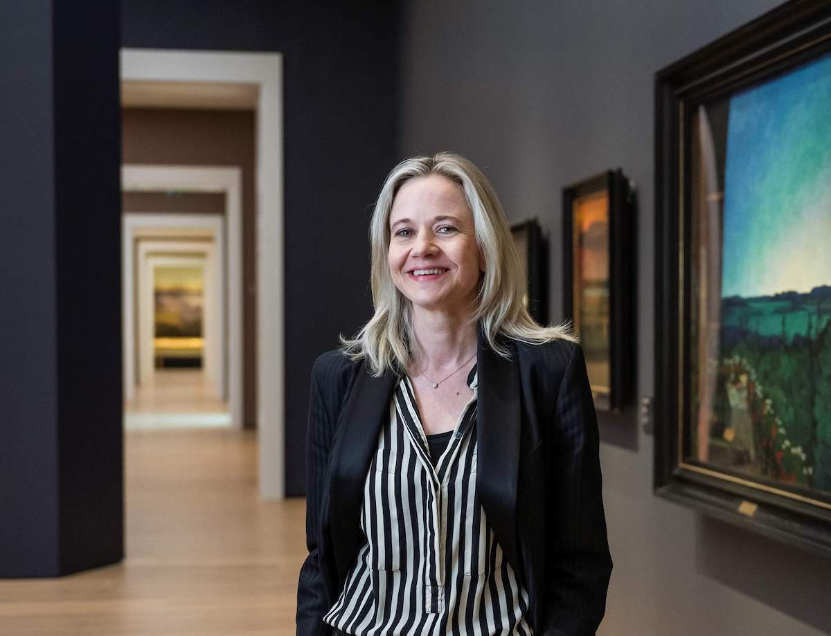 A smiling white woman in museum gallery wearing a blazer and a striped black and white blouse.