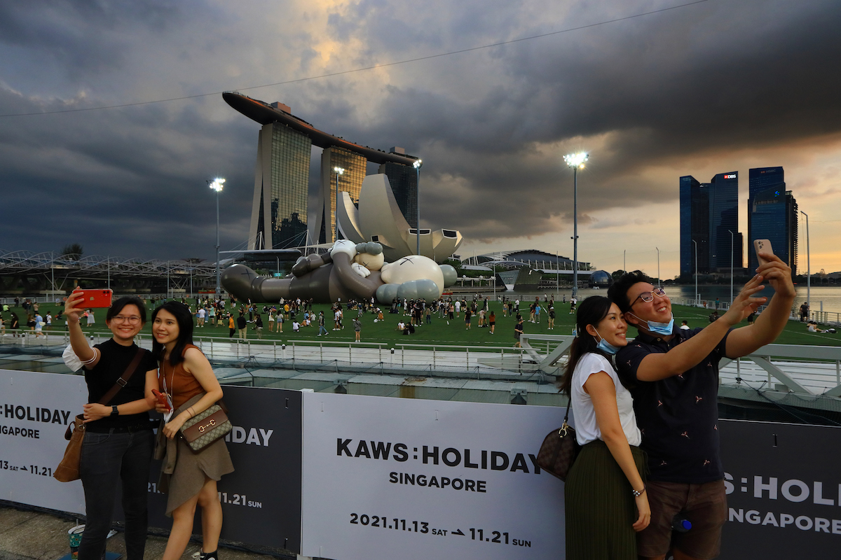 People taking selfies with their phones in front of a giant inflatable sculpture of a figure with Xed out eyes. A dark, cloudy sky looms overhead.