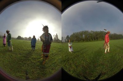 Viewed through two circular apertures, six people in traditional Native attire stand apart from each other in a grassy field.