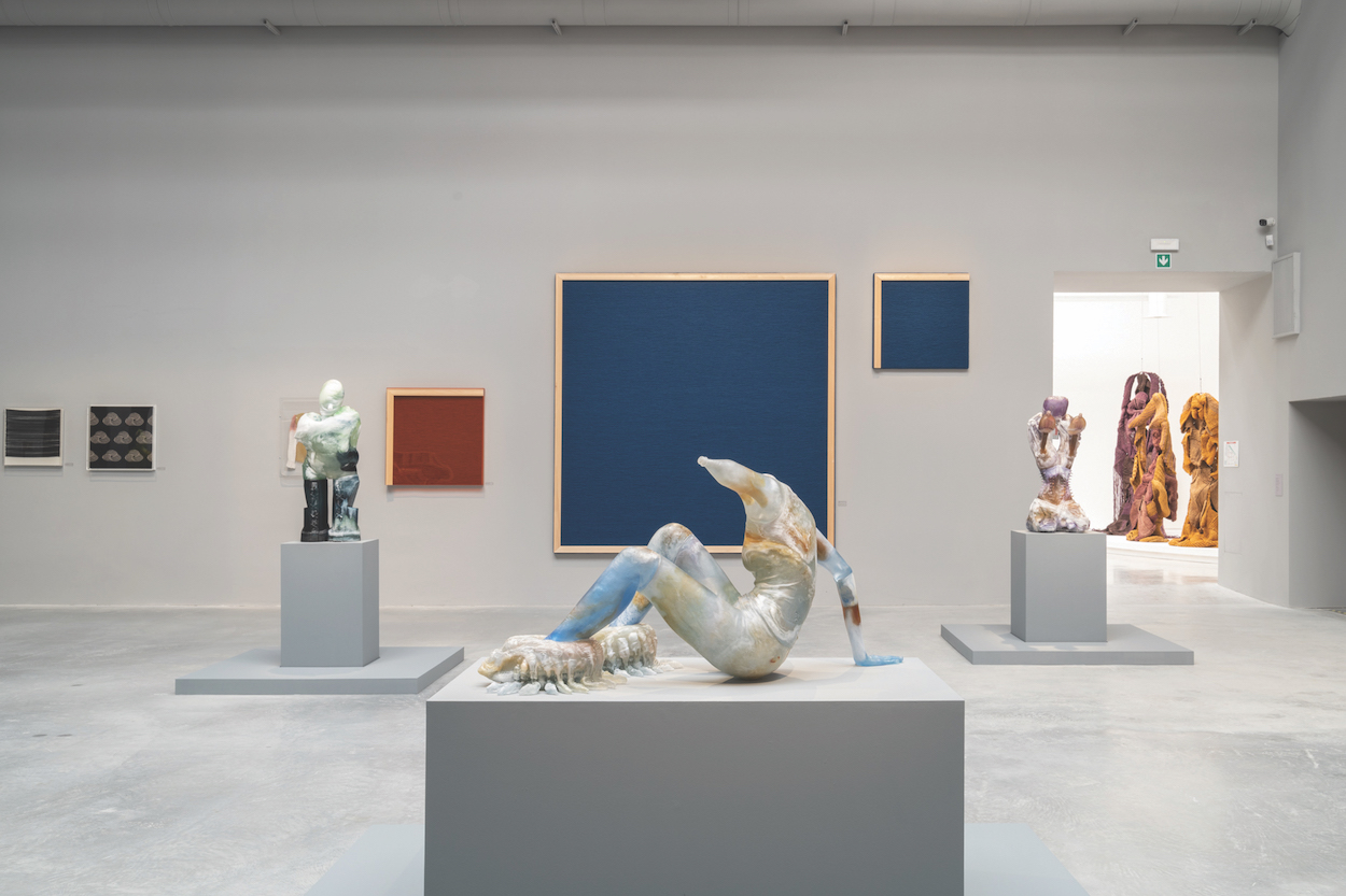 A gallery installation view shows a set of monochromatic wall-hung works at back and, in the foreground, a sculpture of a roughly human form on a pedestal.