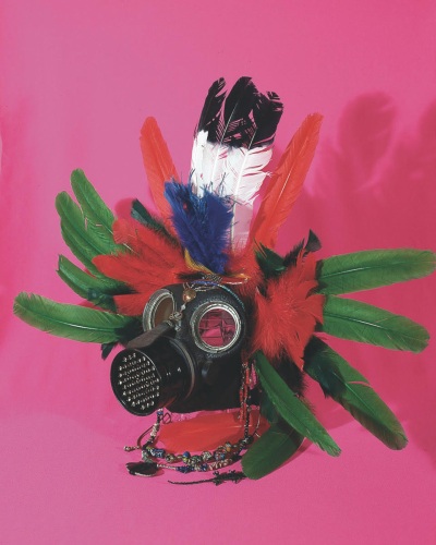 vertical photograph of a gas mask adorned with red and green feathers on a bright pink backdrop