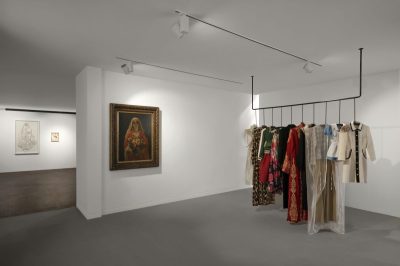A gallery space with a suspended rack of clothes and a painting on the wall, with another gallery visible beyond.