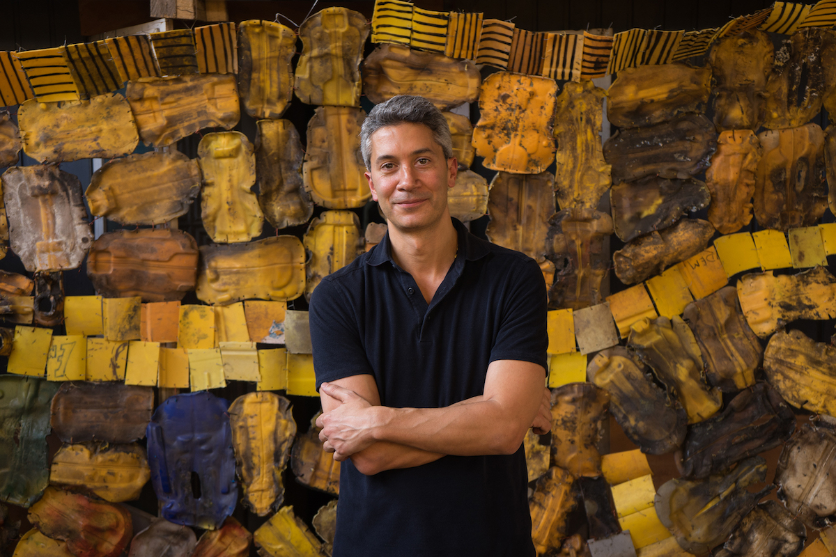 A man wearing a polo shirt standing with his arms crossed before a giant gold-toned installation composed of what appear to be pieces of lumber.