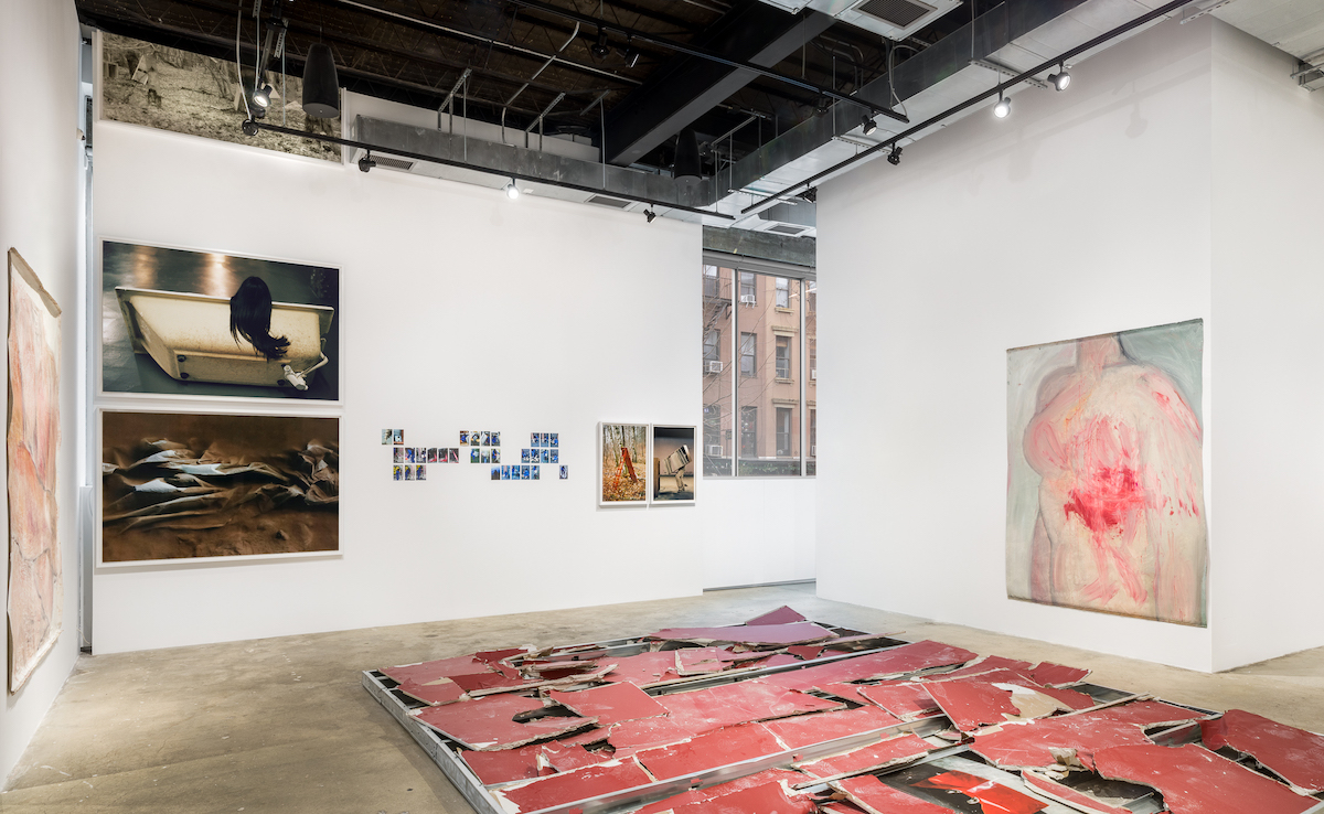 A gallery whose walls are filled with photographs of varying sizes and a painting of a person's chest smeared with a blood-like substance. On the floor lies a torn-up red structure.