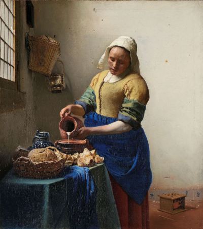 A white woman pouring milk into a vessel sitting atop a table that also has a basket with bread and other assorted items. The woman wears a blue dress and a white hood. A heating device is on the floor, and a basket and a window are on the wall.