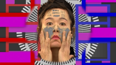 A woman looks into the camera and holds her hands up to her face. She's in the midst of a geometric and striped pattern, some of which also appears on her face.