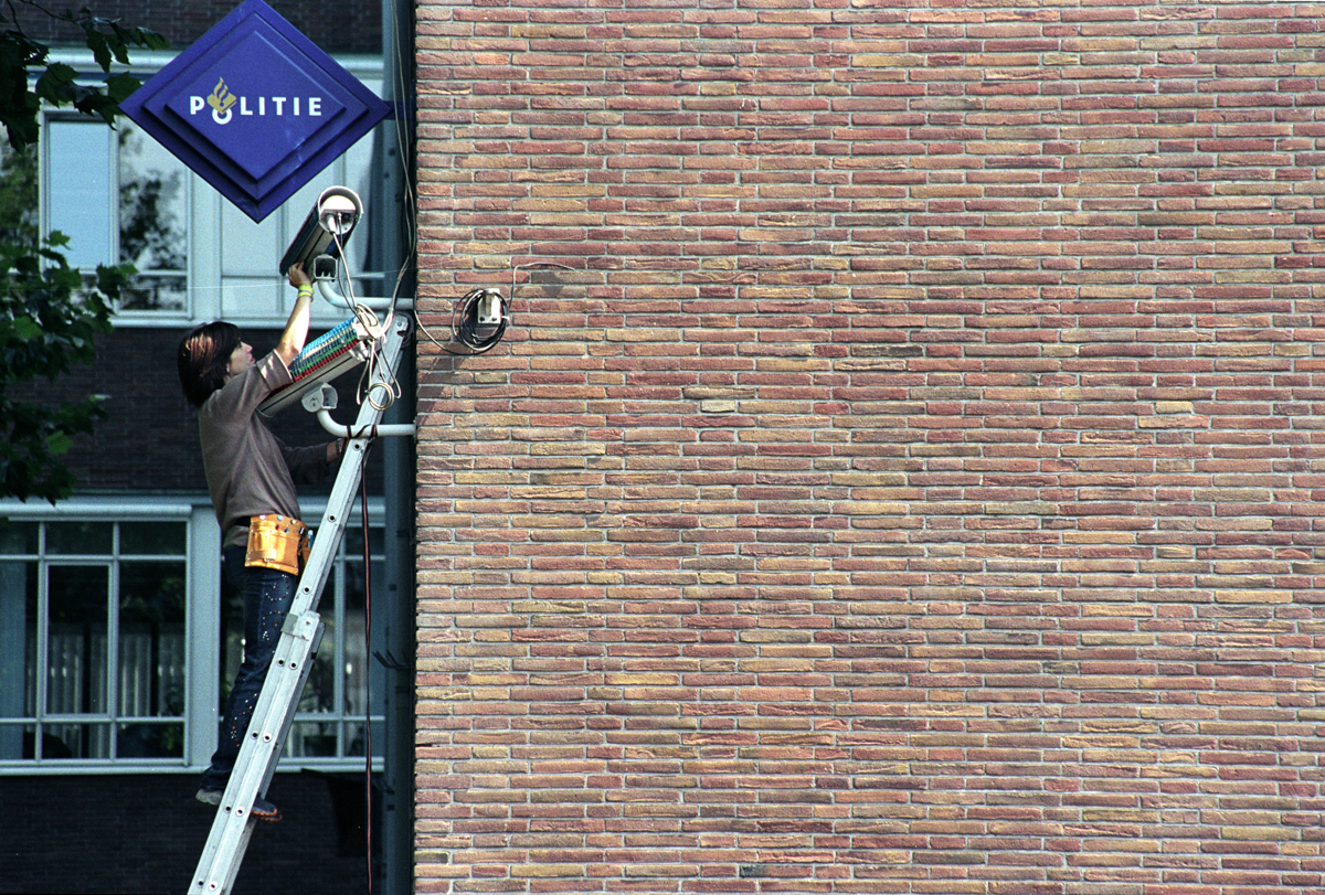 A white woman climbs a ladder leaned against a brick wall. She's attaching rhinestones to security cameras.
