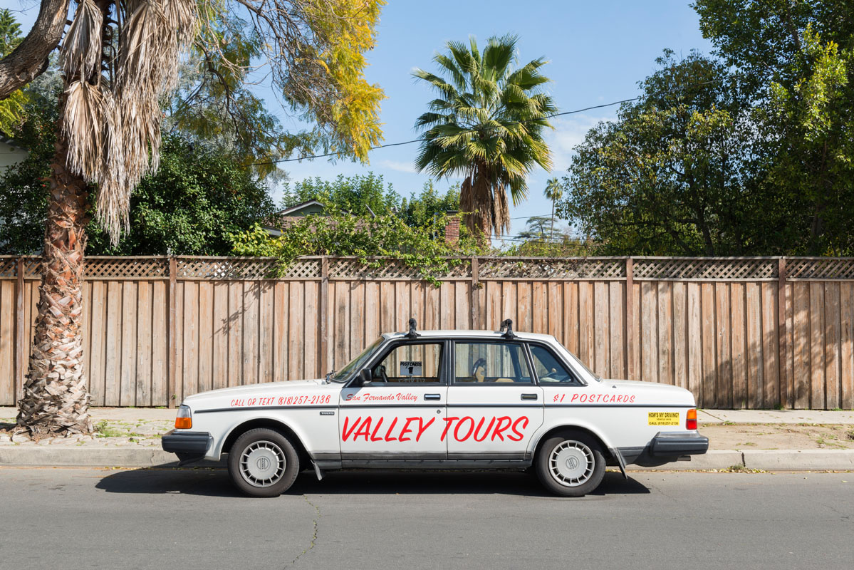 An image of an old-school sedan parked on a sunny street with palm trees in the background. Across the side painted in read reads 'VALLEY TOURS'.