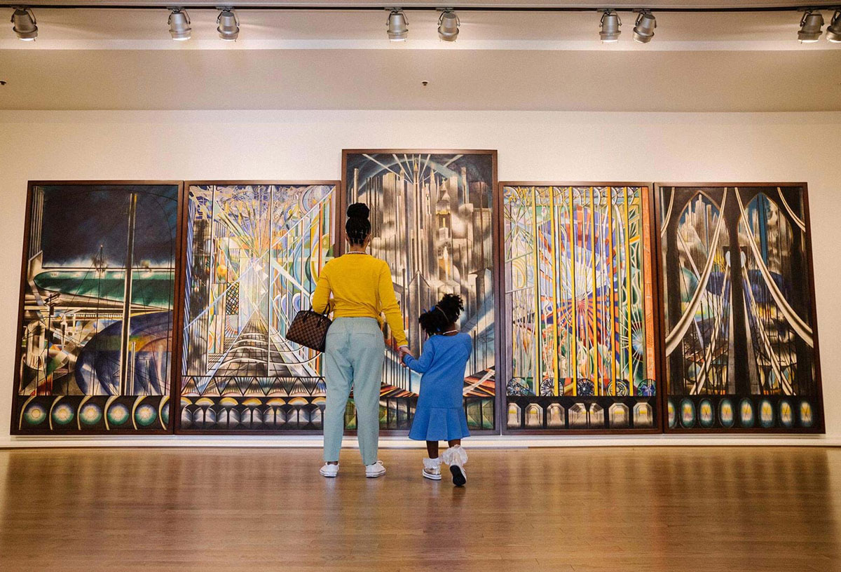 A Black mother and daughter stand before a five-panel artwork.