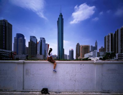 A young girl stills atop a white wall staring at a skyline with a blue-glass skyscraper in the center.
