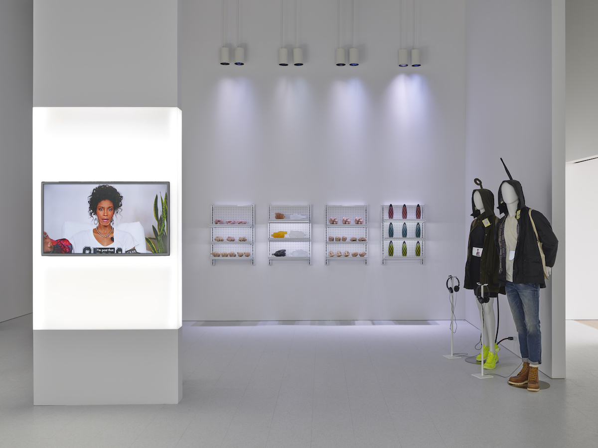 A gallery resembling a sleek white store with spotlighted shelving units, two mannequins wearing coats, and a video screen showing a Black woman speaking.