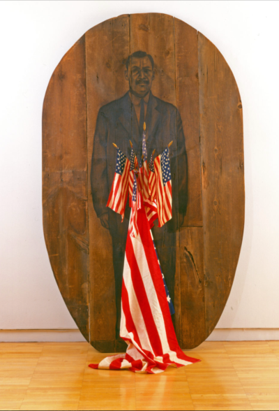 A large oval wood panel bearing a drawn portrait of a man in a suit, with multiple small American flags protruding from the surface and a large one that hangs to the ground.